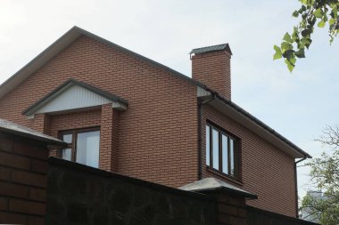 Mavi gökyüzüne karşı çitin arkasında pencereleri olan büyük kahverengi tuğlalı bir ev.