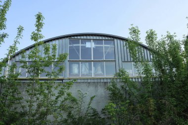 Gökyüzüne bakan büyük pencereli bir fabrikanın gri metal duvarı yeşil bitkilerle çevrili beton bir çitin arkasında.