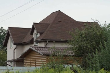 Gökyüzüne karşı yeşil bitki örtüsü içinde gri metal bir çitin arkasında kahverengi kiremitli çatısı olan büyük bir kırsal evin parçası.