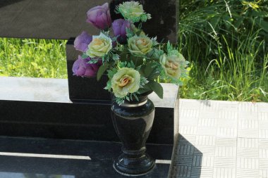  Siyah mermer vazo ve renkli yapay çiçekler mezardaki anıtta duruyor.