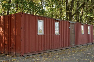 Pencereli büyük kahverengi bir konteynır ve ağaçların önündeki asfaltta duran bir kapı.