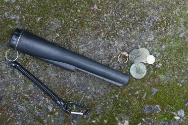 Bir siyah iğne ucu gri bir taşın üzerinde ve yeşil yosun eski paraların yanında ve sokakta altın bir yüzük var.