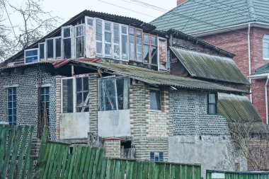 Tuğla ve cam şişelerden yapılmış eski gri çiftlik evinin dışında yeşil bir çitin arkasında birçok pencere var.