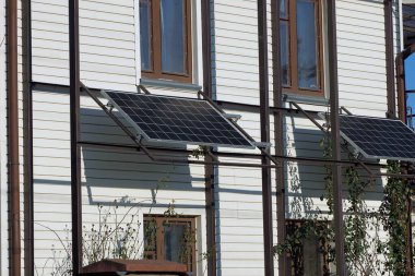 Sokakta pencereleri olan beyaz bir evin beyaz duvarında iki mavi güneş paneli.