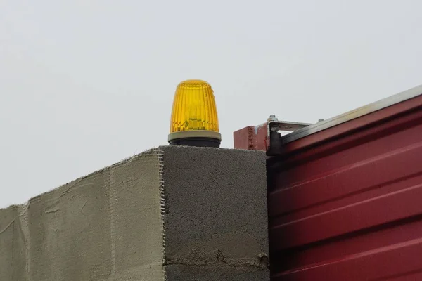one yellow signal lamp on a gray concrete wall of a fence near a red metal gate on the street against the sky