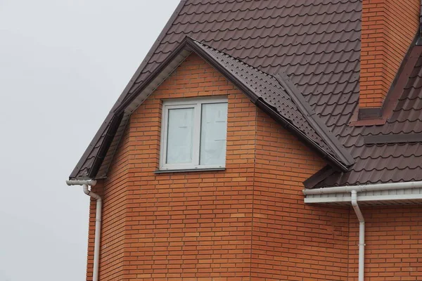 灰色の空に向かって茶色の瓦屋根の下に白い窓のある赤レンガの民家の屋根裏部屋 — ストック写真