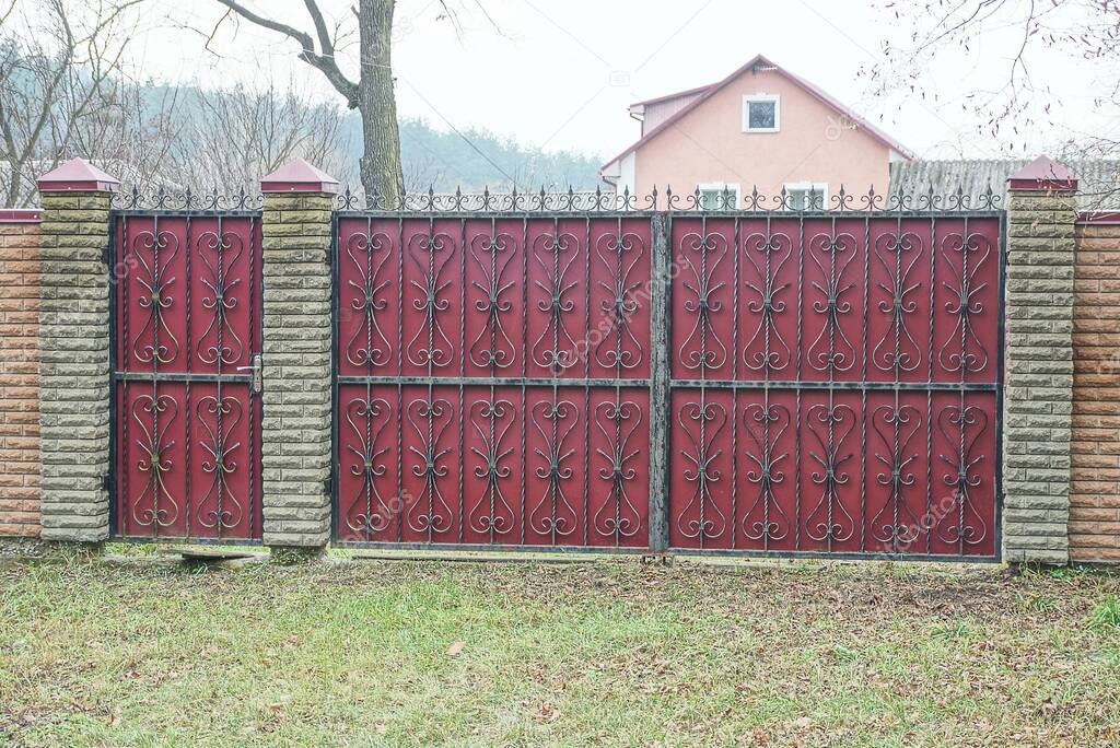 red metal gates with black forged pattern and brown brick fence outside in green grass