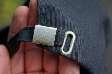 Parmaklar elde, kumaştan yapılmış, koşumlu siyah bir kapak ve paslanmış gri bir metal parçası tutuyor.