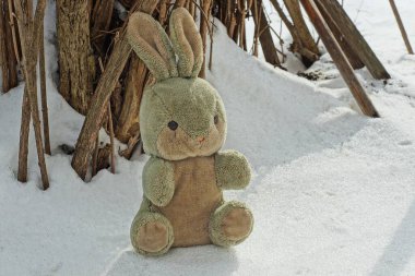 Bir gri oyuncak tavşan, bir kış caddesinde bir çalılığın kahverengi dallarının yanında beyaz karların üzerinde oturur.