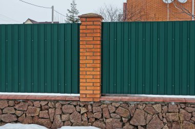 Kırmızı tuğlalardan ve yeşil metalden yapılmış bir çit parçası. Kahverengi taşların üzerine kurulmuş. Kış sokağındaki beyaz karın üzerine kurulmuş.