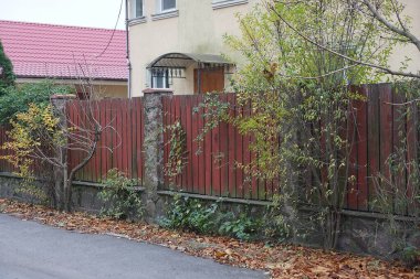 Kırsal bir duvarın bir parçası. Kırmızı tahta ve gri taşlardan yapılmış. Sokakta asfalt bir yolun kenarında yeşil bitki örtüsü var.