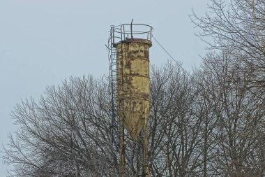 Gri ağaç dalları arasında kahverengi pas kaplı bir merdiveni olan eski sarı demir su kulesi.