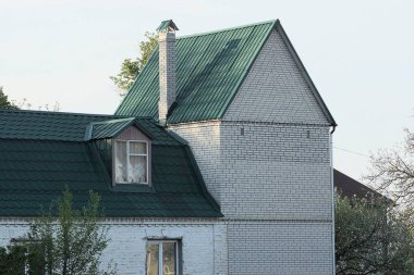 Gri gökyüzüne karşı yeşil kiremitli çatı altında pencereleri olan beyaz tuğlalı bir evin tavan arası.