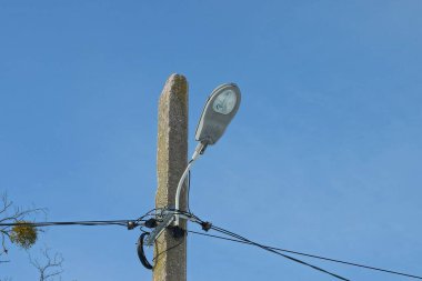  Beton bir direğin üzerinde cam bir ampul ve mavi gökyüzüne karşı siyah elektrik kabloları olan gri demir bir fener.