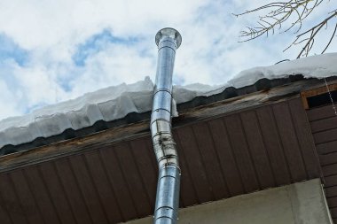 uzun gri baca borusu beyaz karın altında çatıya yakın duvarda gökyüzü ve bulutların arka planına karşı.