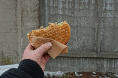 Siyah elbiseli bir el, arka plandaki gri taş ve beyaz kara karşı bir kış caddesinde kağıt bir paketin içinde kahverengi bir sandviç tutuyor.