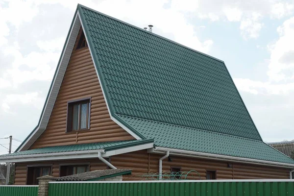 通りの金属製フェンスの後ろの灰色の空に向かって緑のタイル張りの屋根の下に窓のある茶色の民家のファサード — ストック写真