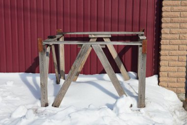 Gri ahşap kalaslardan yapılmış kutunun çerçevesi sokağın kırmızı metal duvarına yakın beyaz kar yığınının içinde duruyor.