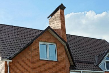 Beyaz pencereli kırmızı tuğla tavan arası ve kahverengi kiremitli çatıdaki büyük baca mavi gökyüzü ve bulutların arka planına karşı.