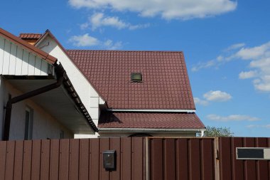 Mavi gökyüzüne karşı metal bir çitin arkasında tek penceresi olan özel bir evin kahverengi kiremitli çatısının bir parçası.