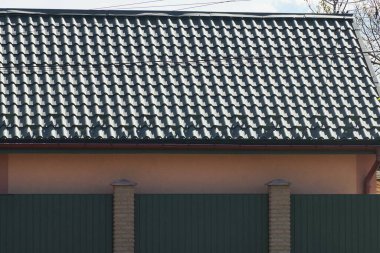 Metal bir duvarın arkasında gri bir gökyüzüne karşı yeşil kiremitli bir çatı.