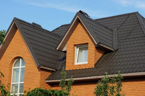 Mavi gökyüzüne karşı kahverengi kiremitli çatı altında pencereleri olan özel kırmızı tuğlalı bir evin tavan arası.