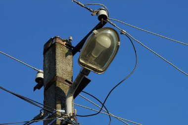  Beton bir direğin üzerinde cam bir ampul ve mavi gökyüzüne karşı siyah elektrik kabloları olan gri bir fener.