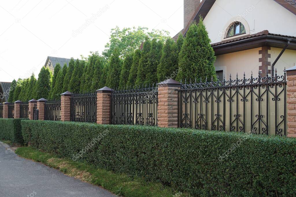 a long fence wall of brown bricks and sharp iron rods outside in green ornamental bushes and a row of long conifers