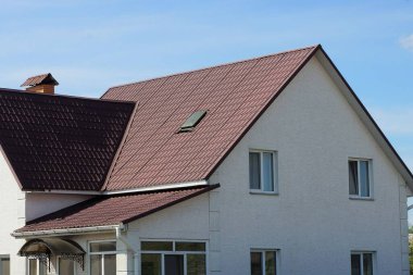 Kahverengi kiremitli çatının altında mavi gökyüzüne karşı pencereleri olan büyük beyaz bir evin cephesi