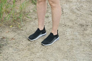 Kumaştan yapılmış siyah beyaz spor ayakkabılı bir kızın bacakları kumsaldaki gri kumların üzerinde duruyor.