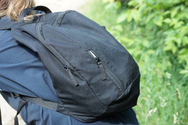 Sokakta mavi gömlekli turist bir kızın sırtındaki kumaştan yapılmış siyah bir sırt çantası yeşil bitkilerin arka planına karşı.