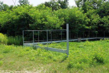 Bitmemiş bir çit, ağaçların arasındaki yeşil çimenlikteki beton bir temelde gri bir metal çerçeveden yapılmıştır.