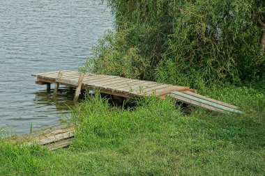 Gri ahşap bir köprü kıyıda, gölün üzerindeki yeşil çimlerde.
