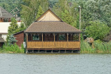 Büyük, ahşap, kahverengi bir ev göl suyunun üzerinde açık bir veranda yeşil bir bitki örtüsü var.