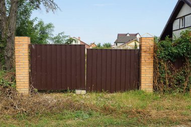 Kahverengi metal kapı ve çitin tuğla duvarının bir kısmı yeşil çimlerde bitki örtüsüyle kaplı.