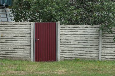 Gri beton bir çit. Sokakta yeşil çimenler ve bitki örtüsü içinde kırmızı bir metal kapı var.