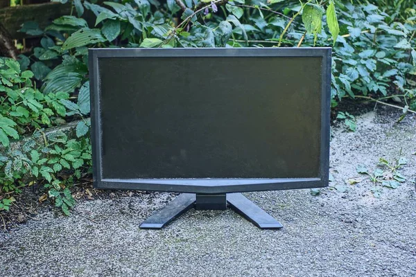 one old black computer monitor stands on gray asphalt near green vegetation on the street