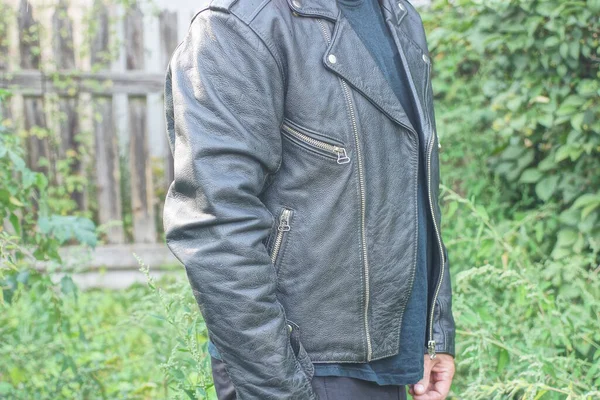 Siyah deri ceketli bir adam yeşil bitki örtüsüne karşı bir yaz caddesinde duruyor.