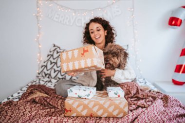 Seçici odaklanma. Güzel bir kadın köpeğiyle yatakta oturuyor ve kameraya Noel hediyesi gösteriyor. O, Noel günü köpeğine sarılırken gülümsüyor. Noel ailesi köpek konsepti