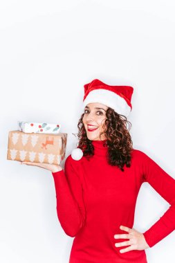 Elinde Noel hediyesi tutan ve kameraya bakan güzel bir kadın. Kırmızı bir kazak ve Noel Baba şapkası giyiyor. Hediye konseptiyle Noel portresi.