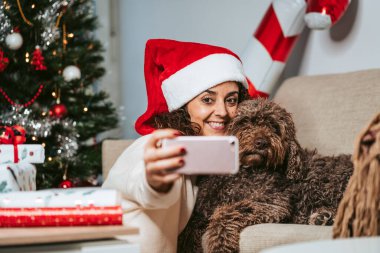 İspanyol su köpeğiyle telefonuyla fotoğraf çeken güzel bir kadın. Noel Baba şapkası ve günlük kıyafetler giyiyor. Evcil hayvanlar ve teknoloji konseptiyle Noel tatili