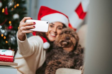 Seçici odaklanma. İspanyol su köpeğiyle telefonuyla fotoğraf çeken esmer bir kadın. Noel Baba şapkası ve günlük kıyafetler giyiyor. Evcil hayvanlar ve teknoloji konseptiyle Noel tatili