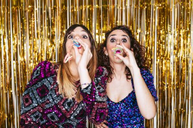 Arkalarında Noel süslemeleri olan iki güzel kadın yeni yıl partisinde kutlama yaparken kameraya flamalar üflemeye hazırlanıyor. Yılbaşı partisi konsepti