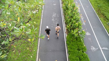 İki koşucu yukarıdan yeşil ve çiçeklerle çevrili asfalt bir yolda görülürler.