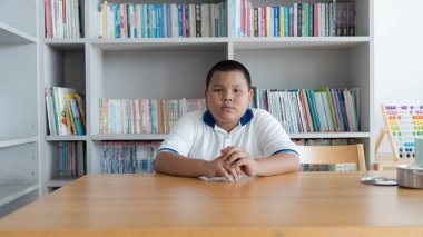 Genç bir çocuk, kitaplar ve öğrenme ortamı ile çevrili, ciddi bir ifadeyle tahta bir masada oturuyor.