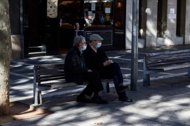 PRAT de LLOBREGAT, İspanya - 24 Ekim 2020: Maskeli iki yaşlı insan, caddedeki ahşap bir bankta oturuyorlar, Covid 19 hakkında konuşuyorlar