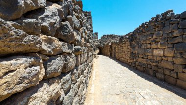 Antik taş duvarlar arasında dar bir patika, açık mavi bir gökyüzünün altındaki kemer yoluna çıkıyor. Bosra antik, Suriye