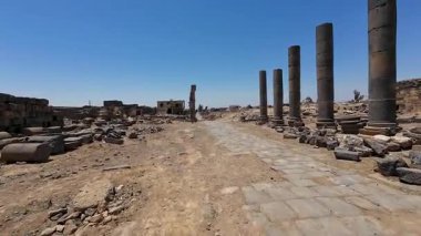 Açık mavi gökyüzünün altında harabeleri ve sütunları olan antik Roma yolu. Bosra antik, Suriye