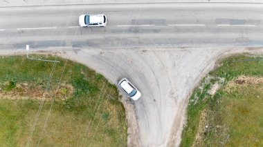 An aerial view of a road intersection with two vehicles, one turning and one driving straight, surrounded by green grass and dirt