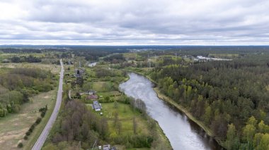 Bereketli yeşil bir arazide dolambaçlı bir nehrin havadan görünüşü dağınık evler ve bulutlu bir gökyüzünün altında bir yol..